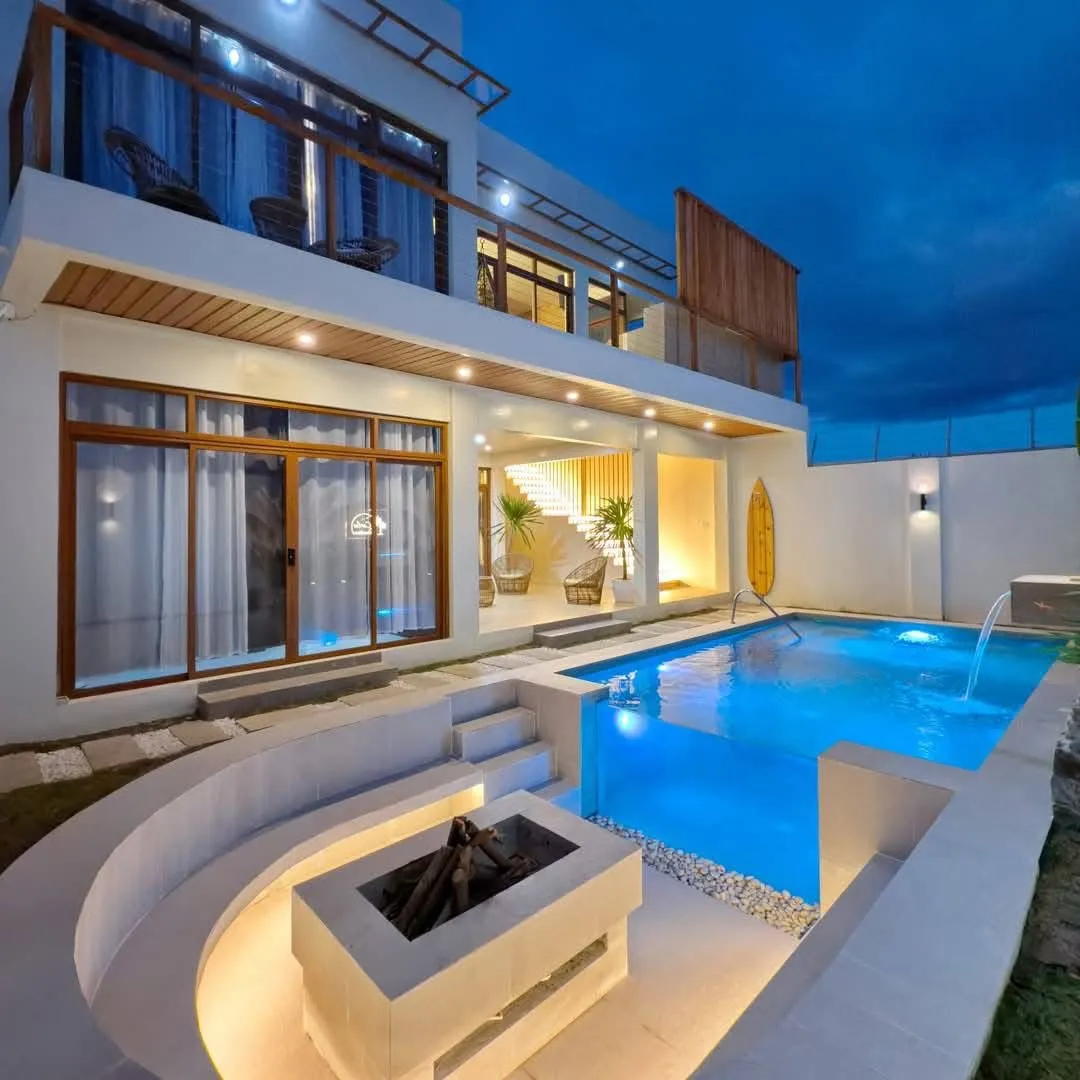 Lit pool at dusk with a glass wall panel, a spout water feature, and a sunken fire pit lounge with built-in curved seating beside it
