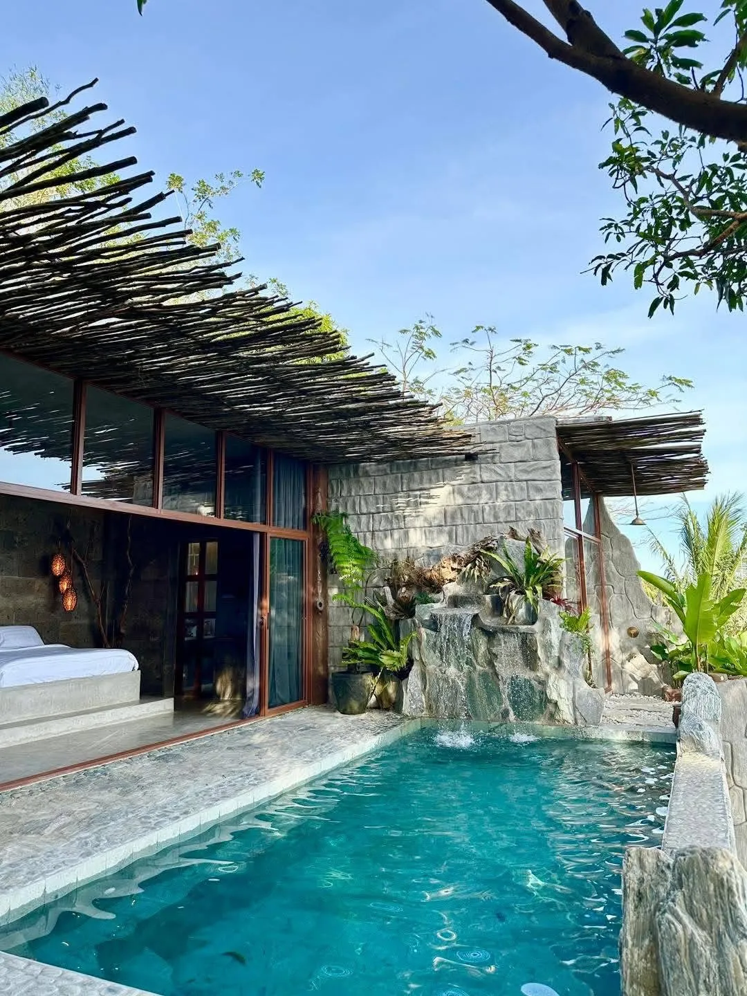 Plunge pool beside a bedroom with a rock waterfall, ferns and staghorn planting, a pebble-set stone deck and a woven branch pergola overhead